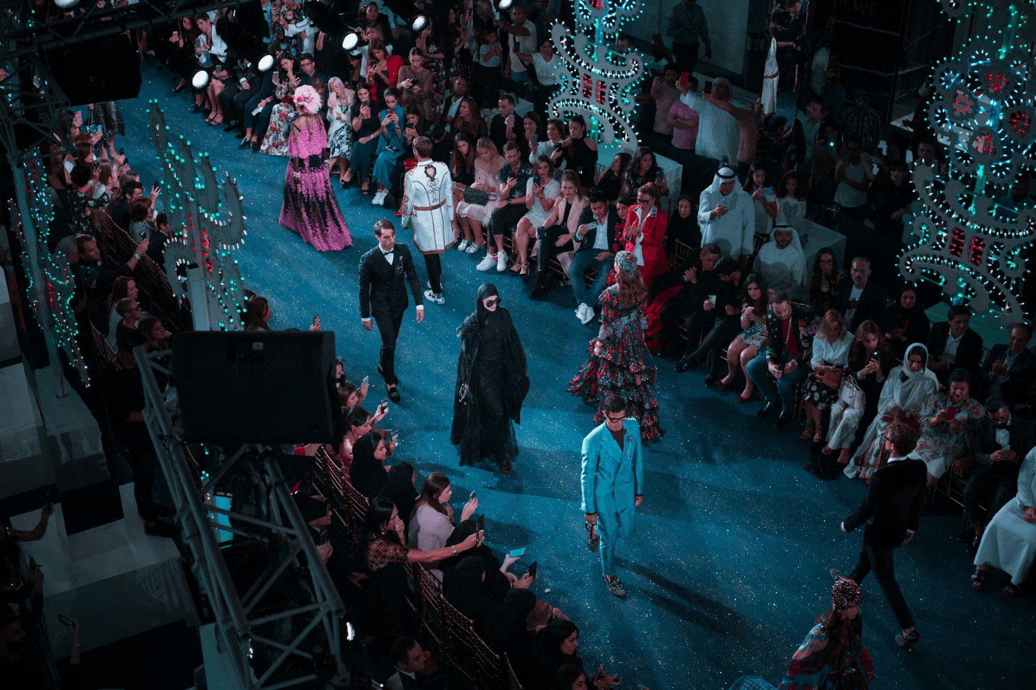 Fashion runway show viewed from above, with models walking the catwalk in elaborate, colorful outfits while an audience watches and takes photos.