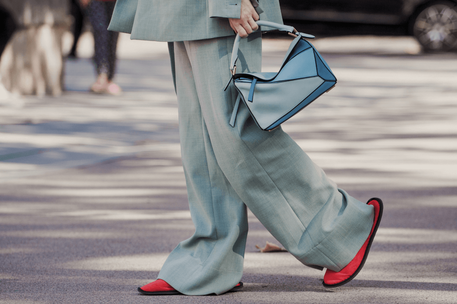 A person wearing an oversized light teal suit walks on the street while carrying a structured blue handbag; the wide pant legs reveal bright red, flat slipper-style shoes.