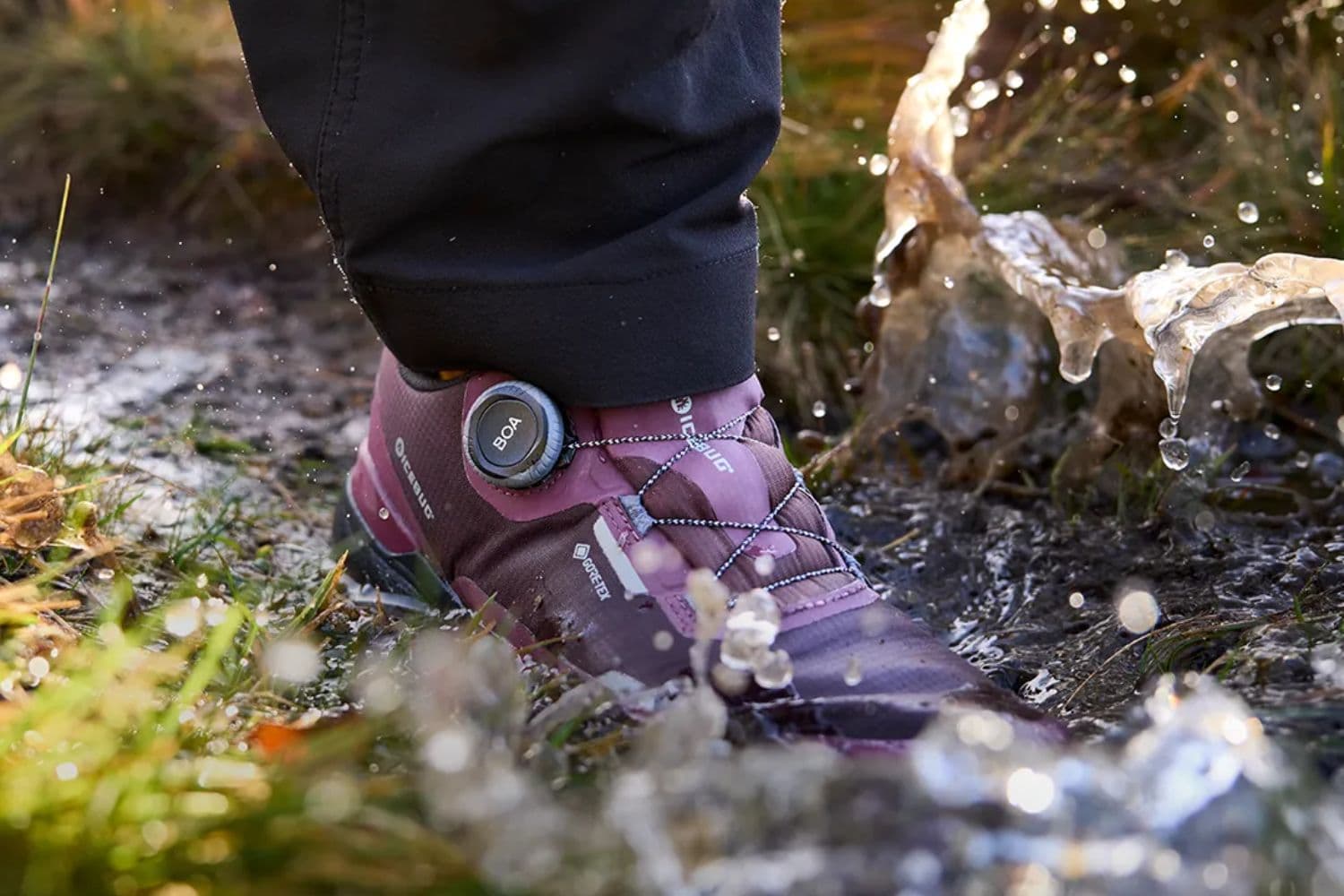 Close-up of a waterproof ICEBUG shoe with a BOA dial lacing system, splashing through a muddy puddle on a trail.